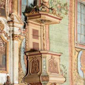 Zdjęcie nr 1: Ambona przyścienna, wisząca, z dostępem od zakrystii dzięki schodom, przez drzwiczki zamontowane w zaplecku. Kosz na planie prostokąta o ściętych narożach, rozszerzającego się ku dołowi, na podstawie w kształcie wałka, z podwieszeniem w formie zbliżonej do stożka o ściętych narożach, wklęsło-wypukłych ściankach, zakończonym szyszką przypominającą tralkę. Parapet w formie belkowania, w miejscu ściętych naroży kosza spływy wolutowe, przechodzące na wałek i podwieszenie, sięgając szyszki, dekorowane płycinami z kampanulami, rocaille'm i zwisami roślinnymi. Ścianki kosza zdobione płycinami w formie kwadratów, o ściętych dolnych narożach, zamkniętych stykającymi się falistymi wolutami z dekoracją roślinną; w polach wypełnione rocaille'm i wiciami roślinnymi. Podwieszenie kosza od frontu dekorowane roślinnym kartuszem z kampanulami w polu, na pozostałych ściankach rocaille'm i ornamentem roślinnym. Zaplecek wąski, ujęty pilastrami dźwigającymi pełne belkowanie. W polu dekorowany dwoma płycinami; z zamontowanymi drzwiczkami. Flankowany uszami utworzonymi z ornamentu roślinnego. Baldachim na planie prostokąta o ściętych narożach; obwiedziony pełnym belkowaniem, wyłamanym w miejscu ściętych naroży i z trzech stron zakończonym szczytami z palmetami. Na baldachimie cztery kabłąki w formie wolut i cęg, stanowiące podstawę dla gorejącego wazonu. Na podniebieniu baldachimu płycina z rzeźbioną gołębicą Ducha Świętego w glorii promienistej. Struktura ambony polichromowana w kolorze beżowym, płyciny i fryzy jasnoczerwonym, spływy wolutowe jasnoniebieskim; profile i ornamentyka złocone i srebrzone. 

