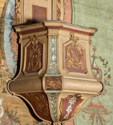 Zdjęcie nr 4: Ambona przyścienna, wisząca, z dostępem od zakrystii dzięki schodom, przez drzwiczki zamontowane w zaplecku. Kosz na planie prostokąta o ściętych narożach, rozszerzającego się ku dołowi, na podstawie w kształcie wałka, z podwieszeniem w formie zbliżonej do stożka o ściętych narożach, wklęsło-wypukłych ściankach, zakończonym szyszką przypominającą tralkę. Parapet w formie belkowania, w miejscu ściętych naroży kosza spływy wolutowe, przechodzące na wałek i podwieszenie, sięgając szyszki, dekorowane płycinami z kampanulami, rocaille&#039;m i zwisami roślinnymi. Ścianki kosza zdobione płycinami w formie kwadratów, o ściętych dolnych narożach, zamkniętych stykającymi się falistymi wolutami z dekoracją roślinną; w polach wypełnione rocaille&#039;m i wiciami roślinnymi. Podwieszenie kosza od frontu dekorowane roślinnym kartuszem z kampanulami w polu, na pozostałych ściankach rocaille&#039;m i ornamentem roślinnym. Zaplecek wąski, ujęty pilastrami dźwigającymi pełne belkowanie. W polu dekorowany dwoma płycinami; z zamontowanymi drzwiczkami. Flankowany uszami utworzonymi z ornamentu roślinnego. Baldachim na planie prostokąta o ściętych narożach; obwiedziony pełnym belkowaniem, wyłamanym w miejscu ściętych naroży i z trzech stron zakończonym szczytami z palmetami. Na baldachimie cztery kabłąki w formie wolut i cęg, stanowiące podstawę dla gorejącego wazonu. Na podniebieniu baldachimu płycina z rzeźbioną gołębicą Ducha Świętego w glorii promienistej. Struktura ambony polichromowana w kolorze beżowym, płyciny i fryzy jasnoczerwonym, spływy wolutowe jasnoniebieskim; profile i ornamentyka złocone i srebrzone. 

