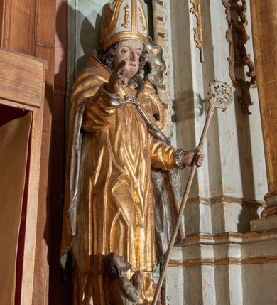 Zdjęcie nr 2: Rzeźba ścięta z tyłu i drążona; ustawiona na niskiej nieregularnej podstawie. Figura ukazana frontalnie, z lekko pochyloną głową, ugiętą prawą nogą, obie ręce ma ugięte, blisko ciała, prawą lekko unosi z dłonią w geście błogosławieństwa, w lewej trzyma pastorał. Twarz ma szeroką, kwadratową, z podkreślonym podbródkiem, o dużych, opadających i półprzymkniętych oczach, sporym, ale wąskim nosie i długich ustach. Włosy sięgające ramion, głęboko rzeźbione w rytmiczne, pojedyncze pukle. Święty ubrany w długą albę, rokietę oraz kapę zarzuconą na ramiona i plecy, zapiętą na piersi klamrą. Na głowie ma infułę, lamowaną taśmami, dekorowaną ornamentem. Przy lewej nodze świętego niewielką postać Piotrowina, klęczącego w skrzyni, ukazanego z profilu, z głową skierowaną w górę i rękami złożonymi w geście modlitwy. Polichromia ciała naturalistyczna, szaty złocone i srebrzone. 