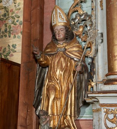 Zdjęcie nr 1: Rzeźba ścięta z tyłu i drążona; ustawiona na niskiej nieregularnej podstawie. Figura ukazana frontalnie, z lekko pochyloną głową, ugiętą prawą nogą, obie ręce ma ugięte, blisko ciała, prawą lekko unosi z dłonią w geście błogosławieństwa, w lewej trzyma pastorał. Twarz ma szeroką, kwadratową, z podkreślonym podbródkiem, o dużych, opadających i półprzymkniętych oczach, sporym, ale wąskim nosie i długich ustach. Włosy sięgające ramion, głęboko rzeźbione w rytmiczne, pojedyncze pukle. Święty ubrany w długą albę, rokietę oraz kapę zarzuconą na ramiona i plecy, zapiętą na piersi klamrą. Na głowie ma infułę, lamowaną taśmami, dekorowaną ornamentem. Przy lewej nodze świętego niewielką postać Piotrowina, klęczącego w skrzyni, ukazanego z profilu, z głową skierowaną w górę i rękami złożonymi w geście modlitwy. Polichromia ciała naturalistyczna, szaty złocone i srebrzone. 