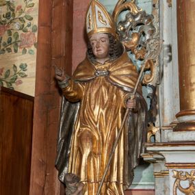 Zdjęcie nr 1: Rzeźba ścięta z tyłu i drążona; ustawiona na niskiej nieregularnej podstawie. Figura ukazana frontalnie, z lekko pochyloną głową, ugiętą prawą nogą, obie ręce ma ugięte, blisko ciała, prawą lekko unosi z dłonią w geście błogosławieństwa, w lewej trzyma pastorał. Twarz ma szeroką, kwadratową, z podkreślonym podbródkiem, o dużych, opadających i półprzymkniętych oczach, sporym, ale wąskim nosie i długich ustach. Włosy sięgające ramion, głęboko rzeźbione w rytmiczne, pojedyncze pukle. Święty ubrany w długą albę, rokietę oraz kapę zarzuconą na ramiona i plecy, zapiętą na piersi klamrą. Na głowie ma infułę, lamowaną taśmami, dekorowaną ornamentem. Przy lewej nodze świętego niewielką postać Piotrowina, klęczącego w skrzyni, ukazanego z profilu, z głową skierowaną w górę i rękami złożonymi w geście modlitwy. Polichromia ciała naturalistyczna, szaty złocone i srebrzone. 