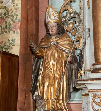 Zdjęcie nr 3: Rzeźba ścięta z tyłu i drążona; ustawiona na niskiej nieregularnej podstawie. Figura ukazana frontalnie, z lekko pochyloną głową, ugiętą prawą nogą, obie ręce ma ugięte, blisko ciała, prawą lekko unosi z dłonią w geście błogosławieństwa, w lewej trzyma pastorał. Twarz ma szeroką, kwadratową, z podkreślonym podbródkiem, o dużych, opadających i półprzymkniętych oczach, sporym, ale wąskim nosie i długich ustach. Włosy sięgające ramion, głęboko rzeźbione w rytmiczne, pojedyncze pukle. Święty ubrany w długą albę, rokietę oraz kapę zarzuconą na ramiona i plecy, zapiętą na piersi klamrą. Na głowie ma infułę, lamowaną taśmami, dekorowaną ornamentem. Przy lewej nodze świętego niewielką postać Piotrowina, klęczącego w skrzyni, ukazanego z profilu, z głową skierowaną w górę i rękami złożonymi w geście modlitwy. Polichromia ciała naturalistyczna, szaty złocone i srebrzone. 