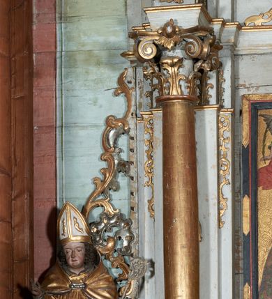 Zdjęcie nr 4: Rzeźba ścięta z tyłu i drążona; ustawiona na niskiej nieregularnej podstawie. Figura ukazana frontalnie, z lekko pochyloną głową, ugiętą prawą nogą, obie ręce ma ugięte, blisko ciała, prawą lekko unosi z dłonią w geście błogosławieństwa, w lewej trzyma pastorał. Twarz ma szeroką, kwadratową, z podkreślonym podbródkiem, o dużych, opadających i półprzymkniętych oczach, sporym, ale wąskim nosie i długich ustach. Włosy sięgające ramion, głęboko rzeźbione w rytmiczne, pojedyncze pukle. Święty ubrany w długą albę, rokietę oraz kapę zarzuconą na ramiona i plecy, zapiętą na piersi klamrą. Na głowie ma infułę, lamowaną taśmami, dekorowaną ornamentem. Przy lewej nodze świętego niewielką postać Piotrowina, klęczącego w skrzyni, ukazanego z profilu, z głową skierowaną w górę i rękami złożonymi w geście modlitwy. Polichromia ciała naturalistyczna, szaty złocone i srebrzone. 