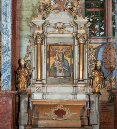 Zdjęcie nr 5: Rzeźba ścięta z tyłu i drążona; ustawiona na niskiej nieregularnej podstawie. Figura ukazana frontalnie, z lekko pochyloną głową, ugiętą prawą nogą, obie ręce ma ugięte, blisko ciała, prawą lekko unosi z dłonią w geście błogosławieństwa, w lewej trzyma pastorał. Twarz ma szeroką, kwadratową, z podkreślonym podbródkiem, o dużych, opadających i półprzymkniętych oczach, sporym, ale wąskim nosie i długich ustach. Włosy sięgające ramion, głęboko rzeźbione w rytmiczne, pojedyncze pukle. Święty ubrany w długą albę, rokietę oraz kapę zarzuconą na ramiona i plecy, zapiętą na piersi klamrą. Na głowie ma infułę, lamowaną taśmami, dekorowaną ornamentem. Przy lewej nodze świętego niewielką postać Piotrowina, klęczącego w skrzyni, ukazanego z profilu, z głową skierowaną w górę i rękami złożonymi w geście modlitwy. Polichromia ciała naturalistyczna, szaty złocone i srebrzone. 