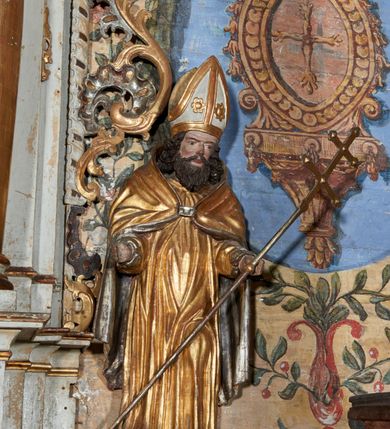 Zdjęcie nr 1: Rzeźba ścięta z tyłu i drążona; ustawiona na niskiej, nieregularnej podstawie. Figura ukazana frontalnie, z lekko ugiętą lewą nogą oraz ugiętymi rękami, rozłożonymi na boki. Twarz szeroka, kwadratowa, o dużych, migdałowatych oczach, z zaznaczonymi powiekami, o lekko zmarszczonych brwiach, małym, wąskim nosie i niewielkich ustach, z zaznaczonymi zmarszczkami i podkreślonymi kośćmi policzkowymi. Włosy sięgające ramion, silnie kręcone w pukle różnej długości. Twarz okolona brodą układającą się w rytmiczne fale, zakończona puklami, z falowanymim długimi wąsami. Ubrany jest w albę i kapę narzuconą na ramiona i plecy, zapiętą na piersi klamrą. Na głowie ma infułę lamowaną taśmami, dekorowaną dwoma rozetami. Polichromią naturalistyczna w partiach ciała, szaty złocone i srebrzone. 