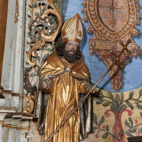 Zdjęcie nr 1: Rzeźba ścięta z tyłu i drążona; ustawiona na niskiej, nieregularnej podstawie. Figura ukazana frontalnie, z lekko ugiętą lewą nogą oraz ugiętymi rękami, rozłożonymi na boki. Twarz szeroka, kwadratowa, o dużych, migdałowatych oczach, z zaznaczonymi powiekami, o lekko zmarszczonych brwiach, małym, wąskim nosie i niewielkich ustach, z zaznaczonymi zmarszczkami i podkreślonymi kośćmi policzkowymi. Włosy sięgające ramion, silnie kręcone w pukle różnej długości. Twarz okolona brodą układającą się w rytmiczne fale, zakończona puklami, z falowanymim długimi wąsami. Ubrany jest w albę i kapę narzuconą na ramiona i plecy, zapiętą na piersi klamrą. Na głowie ma infułę lamowaną taśmami, dekorowaną dwoma rozetami. Polichromią naturalistyczna w partiach ciała, szaty złocone i srebrzone. 