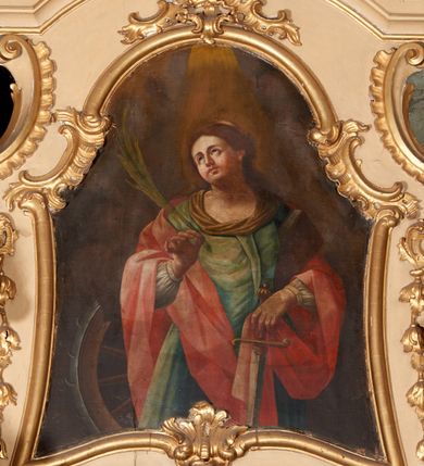 Zdjęcie nr 1: Obraz w formie stojącego prostokąta o wklęsłych bokach, zamkniętego łukiem półkolistym z uskokiem, ujęty profilowaną, złoconą ramą, dekorowaną fragmentami rocaille'u.
W centrum św. Katarzyna ukazana do kolan, frontalnie, z uniesioną głową, skierowaną w prawo. W prawej, ugiętej i uniesionej ręce trzyma liść palmy; lewą, delikatnie opuszczaną przytrzymuje przy boku księgę i dłonią ujmuje rękojeść miecza zwróconego ostrzem w dół. Twarz ma owalną, o pełnych policzkach, o dużych oczach skierowanych w górę, prostym nosie i pełnych ustach. Włosy ma długie, częściowo upięte z tyłu głowy i opadające na plecy. Postać ubrana w białą suknię spodnią, której bufiaste rękawy widoczne są spod płaszcza, jasnozieloną suknię wierzchnią, z zakładką na piersi i brązową tkaniną wokół dekoltu. Na plecy i ramiona ma zarzucony jasnoczerwony, obficie fałdowany płaszcz. Na głowie ma koronę otwartą. Przy jej prawym boku znajduje się ułamane koło obite metalem. Święta ukazana na cieniowanym, brunatnym tle, z wiązką światła padającego z góry na jej głowę.

