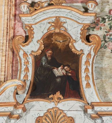 Zdjęcie nr 2: Obraz w formie stojącego prostokąta, o wklęsłych bokach, zamknięty łukiem odcinkowym z uskokiem. W centrum scena ukazująca św. Joachima uczącego Marię przedstawioną jako dziecko. Święty w lewej części obrazu, siedzący w fotelu, zwrócony w trzech czwartych w lewo, z lekko pochyloną głową. Prawą ręką przytrzymuje na kolanach otwartą księgę, lewą zaś wskazuje na tekst w niej zawarty. Twarz ma pociągłą, szczupłą, o starczych rysach, łukowatych brwiach, okrągłych oczach skierowanych w dół, wąskim, garbatym nosie i wąskich ustach; okoloną długą siwą brodą oraz krótkimi włosami. Na głowie ma czerwone nakrycie głowy, ponad nią nimb. Ubrany jest w obfitą, niebieską tunikę, przepasaną w talii oraz zielony płaszcz zarzucony przez lewe przedramię i opadający na kolana. W prawej części obrazu, przed św. Joachimem, ukazana Maria w pozycji stojącej, zwrócona w trzech czwartych w prawo, z pochyloną głową. Obie ręce ma uniesione, prawą dłonią, w geście wskazującym, dotyka księgi. Twarz ma okrągłą, o uniesionych brwiach, okrągłych oczach skierowanych w dół, małym prostym nosie i ustach. Włosy ma długie i brązowe, opadające na plecy. Ubrana jest w długą, czerwoną suknię oraz niebieski płaszcz przerzucony diagonalnie przez prawe ramię. W górnej części obrazu, pośród rozświetlonych obłoków ukazana gołębica Ducha Świętego w glorii promienistej. Postacie ukazane na neutralnym, brązowym tle. 






