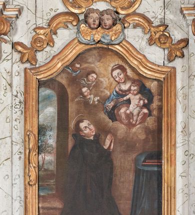 Zdjęcie nr 2: Obraz w formie stojącego prostokąta zamkniętego łukiem falistym nadwieszonym. Przedstawia świętego klęczącego przed Matką Boską z Dzieciątkiem, ukazanych pośród obłoków. W centrum przedstawienia, w dolnej części święty znajduje się w pozycji klęczącej, jest skierowany w trzech czwartych w lewo, z uniesioną głową i rękami złożonymi w geście modlitwy. Twarz ma owalną, o łukowatych brwiach, migdałowatych, dużych oczach skierowanych w górę, długim, prostym nosie, małych ustach i wyeksponowanym podbródku. Głowa okolona krótkimi, brązowymi włosami. Święty ubrany jest w czarną sutannę, z zawieszonym różańcem przy prawym boku, oraz czarny płaszcz ze stojącym kołnierzem, zarzucony na plecy. Nad głową ma nimb. Przed świętym, w prawym dolnym rogu obrazu ukazany stół nakryty niebieską tkaniną, na którym leżą dwie księgi. Ponad nim, w prawym górnym rogu obrazu przedstawiona Matka Boska z Dzieciątkiem, siedząca pośród obłoków, ukazana frontalnie, lekko pochylona w prawo. Lewą ręką przytrzymuje Dzieciątko, w prawej, wyciągniętej w stronę świętego trzyma gałązkę lilii. Twarz ma owalną, o łukowatych brwiach, okrągłych oczach skierowanych w dół, prostym, wąskim nosie, małych ustach i podkreślonym podbródku. Włosy ma długie, jasnobrązowe, upięte z tyłu głowy, z kosmykami opadającymi na ramiona. Ubrana jest w jasnoróżową suknię i niebieski płaszcz zarzucony na ramiona. Wokół głowy Marii nimb promienisty. Dzieciątko ukazane jest w pozycji siedzącej, skierowane w trzech czwartych w prawo. Twarz ma okrągłą, o pełnych policzkach, okrągłych oczach skierowanych w dół, małym nosie i ustach, okoloną krótkimi, jasnymi włosami. W partii bioder ciało ma przepasane białą pieluszką. Po prawej stronie Marii i Dzieciątka trzy uskrzydlone główki anielskie. Scena ukazana jest we wnętrzu, o jasnobrązowych ścianach, z oknem w lewej części obrazu w formie stojącego prostokąta zamkniętego łukiem półkolistym, wychodzącym na pejzaż z drzewem. 


