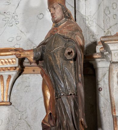 Zdjęcie nr 2: Figura nieopracowana z tyłu; ustawiona na niskiej nieregularnej podstawie. Święty ukazany frontalnie z ugiętą prawą nogą i rękami zgiętymi w łokciach, w tym prawą wyciągniętą w bok z dłonią w geście błogosławieństwa. Twarz ma owalną o prostokątnej żuchwie, niewielkich oczach, krótkim, prostym nosie i małych ustach, okoloną krótkimi włosami skręconymi w pukle. Na głowie ma złoconą infułę z dwoma taśmami. Ubrany jest w albę, rokietę i mucet polichromowane w kolorze ciemnego brązu. Na ramiona ma narzuconą złoconą kapę, zapiętą klamrą w formie czworoliścia, zawiniętą z przodu, na prawej nodze. Na stopie ma but, a na dłoni złoconą rękawiczkę. Polichromia naturalistyczna w odsłoniętych partiach ciała, niektóre elementy stroju złocone (infułą, kapa, rękawiczka).





