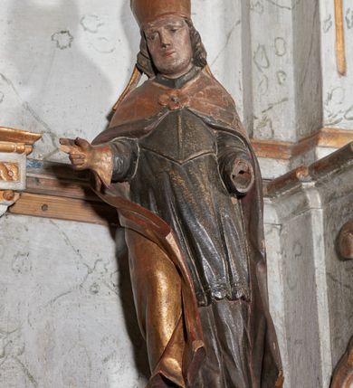 Zdjęcie nr 1: Figura nieopracowana z tyłu; ustawiona na niskiej nieregularnej podstawie. Święty ukazany frontalnie z ugiętą prawą nogą i rękami zgiętymi w łokciach, w tym prawą wyciągniętą w bok z dłonią w geście błogosławieństwa. Twarz ma owalną o prostokątnej żuchwie, niewielkich oczach, krótkim, prostym nosie i małych ustach, okoloną krótkimi włosami skręconymi w pukle. Na głowie ma złoconą infułę z dwoma taśmami. Ubrany jest w albę, rokietę i mucet polichromowane w kolorze ciemnego brązu. Na ramiona ma narzuconą złoconą kapę, zapiętą klamrą w formie czworoliścia, zawiniętą z przodu, na prawej nodze. Na stopie ma but, a na dłoni złoconą rękawiczkę. Polichromia naturalistyczna w odsłoniętych partiach ciała, niektóre elementy stroju złocone (infułą, kapa, rękawiczka).





