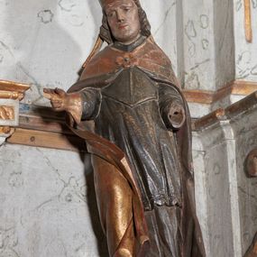Zdjęcie nr 1: Figura nieopracowana z tyłu; ustawiona na niskiej nieregularnej podstawie. Święty ukazany frontalnie z ugiętą prawą nogą i rękami zgiętymi w łokciach, w tym prawą wyciągniętą w bok z dłonią w geście błogosławieństwa. Twarz ma owalną o prostokątnej żuchwie, niewielkich oczach, krótkim, prostym nosie i małych ustach, okoloną krótkimi włosami skręconymi w pukle. Na głowie ma złoconą infułę z dwoma taśmami. Ubrany jest w albę, rokietę i mucet polichromowane w kolorze ciemnego brązu. Na ramiona ma narzuconą złoconą kapę, zapiętą klamrą w formie czworoliścia, zawiniętą z przodu, na prawej nodze. Na stopie ma but, a na dłoni złoconą rękawiczkę. Polichromia naturalistyczna w odsłoniętych partiach ciała, niektóre elementy stroju złocone (infułą, kapa, rękawiczka).





