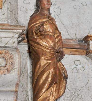 Zdjęcie nr 2: Figura nieopracowana z tyłu; ustawiona na niskiej, nieregularnej podstawie. Święty ukazany frontalnie, prawą, ugiętą rękę wyciąga w bok, lewą, opuszczoną podtrzymuję połę płaszcza. Twarz ma pociągłą, o kwadratowej żuchwie i niskim czole, niewielkich oczach, krótkim, wąskim nosie i małych ustach, okoloną falowanymi włosami opadającymi na ramiona. Ubrany jest w złoconą tunikę przepasaną w talii oraz złocony płaszcz zarzucony na ramiona, zapięty pod szyją klamrą, z połami podtrzymywanymi z przodu postaci. Polichromia ciała naturalistyczna, szaty złocone.


