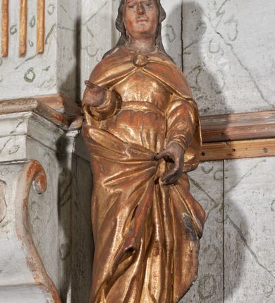 Zdjęcie nr 1: Figura nieopracowana z tyłu; ustawiona na niskiej, nieregularnej podstawie. Święty ukazany frontalnie, prawą, ugiętą rękę wyciąga w bok, lewą, opuszczoną podtrzymuję połę płaszcza. Twarz ma pociągłą, o kwadratowej żuchwie i niskim czole, niewielkich oczach, krótkim, wąskim nosie i małych ustach, okoloną falowanymi włosami opadającymi na ramiona. Ubrany jest w złoconą tunikę przepasaną w talii oraz złocony płaszcz zarzucony na ramiona, zapięty pod szyją klamrą, z połami podtrzymywanymi z przodu postaci. Polichromia ciała naturalistyczna, szaty złocone.


