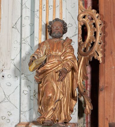 Zdjęcie nr 1: Figura nieopracowana z tyłu; ustawiona na niskiej, nieregularnej podstawie. Święty ukazany frontalnie, z prawą nogą ugiętą, obie ręce wyciąga wprzód, w prawej przytrzymuje księgę. Twarz ma pociągłą, o niewielkich, wąskich oczach, krótkim nosie i małych ustach, okoloną krótkimi, silnie kręconymi włosami i brodą. Ubrany jest w obficie fałdowaną tunikę przepasaną w talii oraz płaszcz zarzucony na lewe ramię i zawinięty z przodu postaci. Polichromia ciała naturalistyczna, szaty złocone. 
