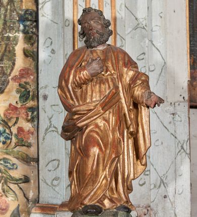 Zdjęcie nr 1: Figura nieopracowana z tyłu; ustawiona na niskiej, nieregularnej podstawie. Święty ukazany frontalnie z prawą nogą ugiętą, lewą rękę wyciąga w bok, prawą kładzie na piersi. Twarz ma pociągłą o niskim czole, niewielkich oczach, krótkim, wąskim nosie i małych ustach, okoloną krótkimi kręconymi włosami i brodą. Ubrany jest w obficie fałdowaną, tunikę przepasaną w talii oraz płaszcz zarzucony na ramiona i zawinięty z przodu postaci. Polichromia ciała naturalistyczna, szaty złocone.




