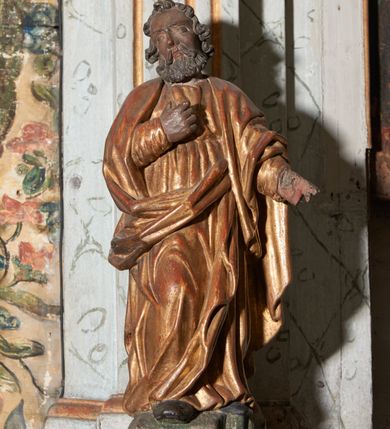 Zdjęcie nr 8: Figura nieopracowana z tyłu; ustawiona na niskiej, nieregularnej podstawie. Święty ukazany frontalnie z prawą nogą ugiętą, lewą rękę wyciąga w bok, prawą kładzie na piersi. Twarz ma pociągłą o niskim czole, niewielkich oczach, krótkim, wąskim nosie i małych ustach, okoloną krótkimi kręconymi włosami i brodą. Ubrany jest w obficie fałdowaną, tunikę przepasaną w talii oraz płaszcz zarzucony na ramiona i zawinięty z przodu postaci. Polichromia ciała naturalistyczna, szaty złocone.




