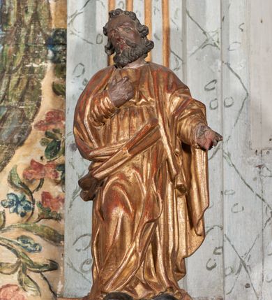Zdjęcie nr 2: Figura nieopracowana z tyłu; ustawiona na niskiej, nieregularnej podstawie. Święty ukazany frontalnie z prawą nogą ugiętą, lewą rękę wyciąga w bok, prawą kładzie na piersi. Twarz ma pociągłą o niskim czole, niewielkich oczach, krótkim, wąskim nosie i małych ustach, okoloną krótkimi kręconymi włosami i brodą. Ubrany jest w obficie fałdowaną, tunikę przepasaną w talii oraz płaszcz zarzucony na ramiona i zawinięty z przodu postaci. Polichromia ciała naturalistyczna, szaty złocone.




