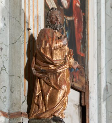 Zdjęcie nr 5: Figura nieopracowana z tyłu; ustawiona na niskiej, nieregularnej podstawie. Święty ukazany frontalnie z prawą nogą ugiętą, lewą rękę wyciąga w bok, prawą kładzie na piersi. Twarz ma pociągłą o niskim czole, niewielkich oczach, krótkim, wąskim nosie i małych ustach, okoloną krótkimi kręconymi włosami i brodą. Ubrany jest w obficie fałdowaną, tunikę przepasaną w talii oraz płaszcz zarzucony na ramiona i zawinięty z przodu postaci. Polichromia ciała naturalistyczna, szaty złocone.




