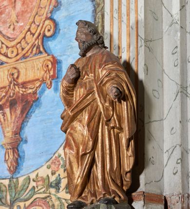 Zdjęcie nr 6: Figura nieopracowana z tyłu; ustawiona na niskiej, nieregularnej podstawie. Święty ukazany frontalnie z prawą nogą ugiętą, lewą rękę wyciąga w bok, prawą kładzie na piersi. Twarz ma pociągłą o niskim czole, niewielkich oczach, krótkim, wąskim nosie i małych ustach, okoloną krótkimi kręconymi włosami i brodą. Ubrany jest w obficie fałdowaną, tunikę przepasaną w talii oraz płaszcz zarzucony na ramiona i zawinięty z przodu postaci. Polichromia ciała naturalistyczna, szaty złocone.




