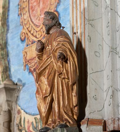 Zdjęcie nr 7: Figura nieopracowana z tyłu; ustawiona na niskiej, nieregularnej podstawie. Święty ukazany frontalnie z prawą nogą ugiętą, lewą rękę wyciąga w bok, prawą kładzie na piersi. Twarz ma pociągłą o niskim czole, niewielkich oczach, krótkim, wąskim nosie i małych ustach, okoloną krótkimi kręconymi włosami i brodą. Ubrany jest w obficie fałdowaną, tunikę przepasaną w talii oraz płaszcz zarzucony na ramiona i zawinięty z przodu postaci. Polichromia ciała naturalistyczna, szaty złocone.




