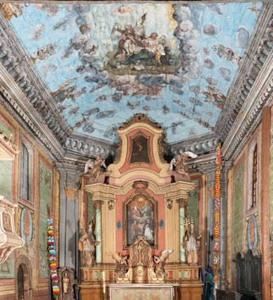 Zdjęcie nr 2: Ściany prezbiterium dekorowane malowanymi zielono-różowymi płycinami w formie stojących prostokątów, ujętymi iluzjonistycznymi ramkami, z malowaną dekoracją w formie kwiatowych festonów w górnej części oraz fantazyjnych dekoracji roślinno-kandelabrowych i wazonów z ornamentem roślinnym w dolnej. Płyciny umieszczone są między oknami ujętymi w malowane prostokątne opaski, pod którymi znajdują się zacheuszki. Na południowej ścianie znajdują się dwa okna, na północnej dwa pozorne okna, z których jedno przesłania ambona. Zacheuszki w formie malowanych migdałowatych medalionów z krzyżami w polach, na iluzjonistycznych wspornikach o klasycyzującej dekoracji, na tle niebieskich owalnych medalionów ujętych u góry wieńcem laurowym i u dołu dekoracją roślinną. Prezbiterium zamknięte trójbocznie, z podkreślonymi polichromią dwiema parami zwielokrotnionych pilastrów, flankujących ołtarz główny. Za ołtarzem, na każdej z trzech ścian analogicznie malowane płyciny. Dolny pas wszystkich ścian prezbiterium opasuje malowany iluzjonistycznie jasnobrązowy i marmoryzowany cokół, dekorowany płycinami w formie leżącego prostokąta i zamknięty profilowaniem. Ściany prezbiterium zamknięte od góry drewnianym gzymsem pokrytym malowanymi iluzjonistycznie elementami profili z kostkowaniem i jajownikiem.
Sklepienie w części prezbiterialnej w formie malowanego nieba, wypełnionego obłokami. Nad gzymsem malowana iluzjonistycznie kamienna balustrada z ośmioma wazonami na cokołach umieszczonych na osiach wydzielających podziały ścian prezbiterium, z motywem stylizowanego biegnącego psa stanowiącym wypełnienie balustrady. Pomiędzy wazonami ukazani jako stojący za balustradą i widoczni w półpostaci aniołowie. Na północnej ścianie anioł trzymający w dłoniach zamknięta koronę, z napisem „PRINCIPATUS” oraz anioł ubrany w zbroję i hełm z białą chorągwią w prawej ręce, podpisany „ARCHANGELI”. Na południowej ścianie anioł ubrany w suknię i płaszcz, który prawą ręką wskazuje niebo, lewą zaś tuli do siebie dziecko, podpisany „ANGELI” oraz anioł ubrany w suknię i płaszcz, który lewą rękę kładzie na piersi, w prawej trzyma miecz skierowany ostrzem w dół, podpisany „DOMINATIONES”. W centralnej części sklepienia ujęta obłokami z malowanym u dołu aniołem scena ekstazy św. Marii Magdaleny. Ukazuje ona omdlewającą na skałach świętą, której towarzyszą aniołowie. Maria Magdalena przedstawiona z profilu, jako siedząca, odchylona w tył, z opadającą głową i rękami. Ubrana w biało-niebieską suknię odsłaniającą ramiona. Włosy ma długie, opadające na plecy i pierś. Nad głową nimb. Za nią i obok niej siedzą dwaj aniołowie, który podtrzymują jej opadające ciało. Przy prawym boku świętej znajduje się krucyfiks, natomiast w dolnej części sceny, na ziemi leżą: czaszka, dyscyplina, otwarta księga i puszka na wonności. W górnej części sceny ukazane rozświetlone niebo, z którego wyłania się wiązka światła, a wokół ukazane są trzy uskrzydlone główki anielskie. Na ścianie zachodniej prezbiterium, nad łukiem tęczowym napis: „Roku Pańskiego 1565 kościół ten zbudowany przez wielmożnego Pana Spytka Jordana, poświęcony przez najjaśniejszego i przewielebnego Pana Tomasza Oborskiego, Biskupa Loadycejskiego, Syfragana Krakowskiego w Roku Pańskim 1634 w dniu 5 czerwca. Odnowiony całkowicie i ozdobiony ołtarzami wraz z organami i chórem przez plebana w Roku pańskim 1778. Odmalowany zaś kosztem tegoż plebana w Roku Pańskim 1802. A[ndrzej] A[ntałkiewicz]”. Za ołtarzem głównym drugi napis: „Restau/rata Hoc / Ecclesia / AD 1777 / Finita 23 / Septembris”.
W korpusie nawowym polichromia o analogicznej formie. Na ścianach północnej i południowej malowane płyciny pomiędzy oknami, okna w malowanych opaskach z zacheuszkami w formie medalionów poniżej. Ściana tęczowa zdobiona malowanymi płycinami, łuk ujęty malowanym en grisaille owocowo-kwiatowym festonem, opadającym na wsporniki z malowanymi iluzjonistycznie elementami architektonicznymi. Ściana zachodnia w dolnej partii malowana analogicznie do ścian bocznych. Natomiast w górnej partii, od poziomu parapetu chóru muzycznego malowana iluzjonistycznie architektura w postaci opilastrowanego muru kamiennego i arkady pośrodku z apsydą o podziałach pilastrowych z malowanymi oknami, na której tle umieszczony jest organ. Zaskrzynienia z malowanymi prostokątnymi płycinami. Filary podtrzymujące zaskrzynienia malowane na sposób imitujący marmoryzację. Podpory podtrzymujące drewniany profilowany gzyms pokryty malowanymi iluzjonistycznie elementami profili z kostkowaniem i jajownikiem. Powyżej sklepienie z malowaną iluzjonistycznie balustradą, z trzech stron (północnej, wschodniej i południowej), analogiczną jak w prezbiterium, z wazonami na cokołach umieszczonymi na osiach podpór i z postaciami. Na ścianie północnej anioł trzymający w dłoniach koronę cierniową z podpisem „VIRTUTES”, anioł trzymający księgę i berło, z podpisem „SERAFIN” oraz anioł trzymający berło, bez podpisu. Na ścianie południowej anioł trzymający buławę, z napisem „POTESTATES”, anioł trzymający miecz skierowany w górę, z napisem „CHERUBIN” i wizerunek św. Franciszka serafickiego podczas stygmatyzacji. Na sklepieniu nawy przedstawione niebo wypełnione obłokami, pośrodku scena Koronacji Najświętszej Marii Panny, zamknięta owalem i otoczona obłokami. Scena ukazuje Chrystusa i Boga Ojca tronujących pośród obłoków z gołębicą Ducha Świętego w glorii promienistej unoszącą się nad nimi. Obaj trzymają koronę nad głową klęczącej poniżej Marii, której nogi spoczywają na globie i sierpie księżyca, a która stopami depcze węża. Grupa otoczona uskrzydlonymi główkami anielskimi, dwoma puttami i aniołem trzymającym w dłoni lilię. 
