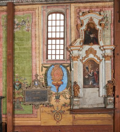 Zdjęcie nr 6: Ściany prezbiterium dekorowane malowanymi zielono-różowymi płycinami w formie stojących prostokątów, ujętymi iluzjonistycznymi ramkami, z malowaną dekoracją w formie kwiatowych festonów w górnej części oraz fantazyjnych dekoracji roślinno-kandelabrowych i wazonów z ornamentem roślinnym w dolnej. Płyciny umieszczone są między oknami ujętymi w malowane prostokątne opaski, pod którymi znajdują się zacheuszki. Na południowej ścianie znajdują się dwa okna, na północnej dwa pozorne okna, z których jedno przesłania ambona. Zacheuszki w formie malowanych migdałowatych medalionów z krzyżami w polach, na iluzjonistycznych wspornikach o klasycyzującej dekoracji, na tle niebieskich owalnych medalionów ujętych u góry wieńcem laurowym i u dołu dekoracją roślinną. Prezbiterium zamknięte trójbocznie, z podkreślonymi polichromią dwiema parami zwielokrotnionych pilastrów, flankujących ołtarz główny. Za ołtarzem, na każdej z trzech ścian analogicznie malowane płyciny. Dolny pas wszystkich ścian prezbiterium opasuje malowany iluzjonistycznie jasnobrązowy i marmoryzowany cokół, dekorowany płycinami w formie leżącego prostokąta i zamknięty profilowaniem. Ściany prezbiterium zamknięte od góry drewnianym gzymsem pokrytym malowanymi iluzjonistycznie elementami profili z kostkowaniem i jajownikiem.
Sklepienie w części prezbiterialnej w formie malowanego nieba, wypełnionego obłokami. Nad gzymsem malowana iluzjonistycznie kamienna balustrada z ośmioma wazonami na cokołach umieszczonych na osiach wydzielających podziały ścian prezbiterium, z motywem stylizowanego biegnącego psa stanowiącym wypełnienie balustrady. Pomiędzy wazonami ukazani jako stojący za balustradą i widoczni w półpostaci aniołowie. Na północnej ścianie anioł trzymający w dłoniach zamknięta koronę, z napisem „PRINCIPATUS” oraz anioł ubrany w zbroję i hełm z białą chorągwią w prawej ręce, podpisany „ARCHANGELI”. Na południowej ścianie anioł ubrany w suknię i płaszcz, który prawą ręką wskazuje niebo, lewą zaś tuli do siebie dziecko, podpisany „ANGELI” oraz anioł ubrany w suknię i płaszcz, który lewą rękę kładzie na piersi, w prawej trzyma miecz skierowany ostrzem w dół, podpisany „DOMINATIONES”. W centralnej części sklepienia ujęta obłokami z malowanym u dołu aniołem scena ekstazy św. Marii Magdaleny. Ukazuje ona omdlewającą na skałach świętą, której towarzyszą aniołowie. Maria Magdalena przedstawiona z profilu, jako siedząca, odchylona w tył, z opadającą głową i rękami. Ubrana w biało-niebieską suknię odsłaniającą ramiona. Włosy ma długie, opadające na plecy i pierś. Nad głową nimb. Za nią i obok niej siedzą dwaj aniołowie, który podtrzymują jej opadające ciało. Przy prawym boku świętej znajduje się krucyfiks, natomiast w dolnej części sceny, na ziemi leżą: czaszka, dyscyplina, otwarta księga i puszka na wonności. W górnej części sceny ukazane rozświetlone niebo, z którego wyłania się wiązka światła, a wokół ukazane są trzy uskrzydlone główki anielskie. Na ścianie zachodniej prezbiterium, nad łukiem tęczowym napis: „Roku Pańskiego 1565 kościół ten zbudowany przez wielmożnego Pana Spytka Jordana, poświęcony przez najjaśniejszego i przewielebnego Pana Tomasza Oborskiego, Biskupa Loadycejskiego, Syfragana Krakowskiego w Roku Pańskim 1634 w dniu 5 czerwca. Odnowiony całkowicie i ozdobiony ołtarzami wraz z organami i chórem przez plebana w Roku pańskim 1778. Odmalowany zaś kosztem tegoż plebana w Roku Pańskim 1802. A[ndrzej] A[ntałkiewicz]”. Za ołtarzem głównym drugi napis: „Restau/rata Hoc / Ecclesia / AD 1777 / Finita 23 / Septembris”.
W korpusie nawowym polichromia o analogicznej formie. Na ścianach północnej i południowej malowane płyciny pomiędzy oknami, okna w malowanych opaskach z zacheuszkami w formie medalionów poniżej. Ściana tęczowa zdobiona malowanymi płycinami, łuk ujęty malowanym en grisaille owocowo-kwiatowym festonem, opadającym na wsporniki z malowanymi iluzjonistycznie elementami architektonicznymi. Ściana zachodnia w dolnej partii malowana analogicznie do ścian bocznych. Natomiast w górnej partii, od poziomu parapetu chóru muzycznego malowana iluzjonistycznie architektura w postaci opilastrowanego muru kamiennego i arkady pośrodku z apsydą o podziałach pilastrowych z malowanymi oknami, na której tle umieszczony jest organ. Zaskrzynienia z malowanymi prostokątnymi płycinami. Filary podtrzymujące zaskrzynienia malowane na sposób imitujący marmoryzację. Podpory podtrzymujące drewniany profilowany gzyms pokryty malowanymi iluzjonistycznie elementami profili z kostkowaniem i jajownikiem. Powyżej sklepienie z malowaną iluzjonistycznie balustradą, z trzech stron (północnej, wschodniej i południowej), analogiczną jak w prezbiterium, z wazonami na cokołach umieszczonymi na osiach podpór i z postaciami. Na ścianie północnej anioł trzymający w dłoniach koronę cierniową z podpisem „VIRTUTES”, anioł trzymający księgę i berło, z podpisem „SERAFIN” oraz anioł trzymający berło, bez podpisu. Na ścianie południowej anioł trzymający buławę, z napisem „POTESTATES”, anioł trzymający miecz skierowany w górę, z napisem „CHERUBIN” i wizerunek św. Franciszka serafickiego podczas stygmatyzacji. Na sklepieniu nawy przedstawione niebo wypełnione obłokami, pośrodku scena Koronacji Najświętszej Marii Panny, zamknięta owalem i otoczona obłokami. Scena ukazuje Chrystusa i Boga Ojca tronujących pośród obłoków z gołębicą Ducha Świętego w glorii promienistej unoszącą się nad nimi. Obaj trzymają koronę nad głową klęczącej poniżej Marii, której nogi spoczywają na globie i sierpie księżyca, a która stopami depcze węża. Grupa otoczona uskrzydlonymi główkami anielskimi, dwoma puttami i aniołem trzymającym w dłoni lilię. 
