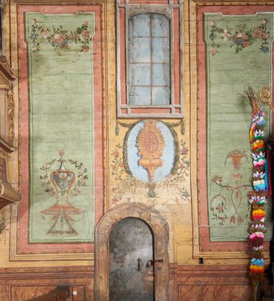 Zdjęcie nr 7: Ściany prezbiterium dekorowane malowanymi zielono-różowymi płycinami w formie stojących prostokątów, ujętymi iluzjonistycznymi ramkami, z malowaną dekoracją w formie kwiatowych festonów w górnej części oraz fantazyjnych dekoracji roślinno-kandelabrowych i wazonów z ornamentem roślinnym w dolnej. Płyciny umieszczone są między oknami ujętymi w malowane prostokątne opaski, pod którymi znajdują się zacheuszki. Na południowej ścianie znajdują się dwa okna, na północnej dwa pozorne okna, z których jedno przesłania ambona. Zacheuszki w formie malowanych migdałowatych medalionów z krzyżami w polach, na iluzjonistycznych wspornikach o klasycyzującej dekoracji, na tle niebieskich owalnych medalionów ujętych u góry wieńcem laurowym i u dołu dekoracją roślinną. Prezbiterium zamknięte trójbocznie, z podkreślonymi polichromią dwiema parami zwielokrotnionych pilastrów, flankujących ołtarz główny. Za ołtarzem, na każdej z trzech ścian analogicznie malowane płyciny. Dolny pas wszystkich ścian prezbiterium opasuje malowany iluzjonistycznie jasnobrązowy i marmoryzowany cokół, dekorowany płycinami w formie leżącego prostokąta i zamknięty profilowaniem. Ściany prezbiterium zamknięte od góry drewnianym gzymsem pokrytym malowanymi iluzjonistycznie elementami profili z kostkowaniem i jajownikiem.
Sklepienie w części prezbiterialnej w formie malowanego nieba, wypełnionego obłokami. Nad gzymsem malowana iluzjonistycznie kamienna balustrada z ośmioma wazonami na cokołach umieszczonych na osiach wydzielających podziały ścian prezbiterium, z motywem stylizowanego biegnącego psa stanowiącym wypełnienie balustrady. Pomiędzy wazonami ukazani jako stojący za balustradą i widoczni w półpostaci aniołowie. Na północnej ścianie anioł trzymający w dłoniach zamknięta koronę, z napisem „PRINCIPATUS” oraz anioł ubrany w zbroję i hełm z białą chorągwią w prawej ręce, podpisany „ARCHANGELI”. Na południowej ścianie anioł ubrany w suknię i płaszcz, który prawą ręką wskazuje niebo, lewą zaś tuli do siebie dziecko, podpisany „ANGELI” oraz anioł ubrany w suknię i płaszcz, który lewą rękę kładzie na piersi, w prawej trzyma miecz skierowany ostrzem w dół, podpisany „DOMINATIONES”. W centralnej części sklepienia ujęta obłokami z malowanym u dołu aniołem scena ekstazy św. Marii Magdaleny. Ukazuje ona omdlewającą na skałach świętą, której towarzyszą aniołowie. Maria Magdalena przedstawiona z profilu, jako siedząca, odchylona w tył, z opadającą głową i rękami. Ubrana w biało-niebieską suknię odsłaniającą ramiona. Włosy ma długie, opadające na plecy i pierś. Nad głową nimb. Za nią i obok niej siedzą dwaj aniołowie, który podtrzymują jej opadające ciało. Przy prawym boku świętej znajduje się krucyfiks, natomiast w dolnej części sceny, na ziemi leżą: czaszka, dyscyplina, otwarta księga i puszka na wonności. W górnej części sceny ukazane rozświetlone niebo, z którego wyłania się wiązka światła, a wokół ukazane są trzy uskrzydlone główki anielskie. Na ścianie zachodniej prezbiterium, nad łukiem tęczowym napis: „Roku Pańskiego 1565 kościół ten zbudowany przez wielmożnego Pana Spytka Jordana, poświęcony przez najjaśniejszego i przewielebnego Pana Tomasza Oborskiego, Biskupa Loadycejskiego, Syfragana Krakowskiego w Roku Pańskim 1634 w dniu 5 czerwca. Odnowiony całkowicie i ozdobiony ołtarzami wraz z organami i chórem przez plebana w Roku pańskim 1778. Odmalowany zaś kosztem tegoż plebana w Roku Pańskim 1802. A[ndrzej] A[ntałkiewicz]”. Za ołtarzem głównym drugi napis: „Restau/rata Hoc / Ecclesia / AD 1777 / Finita 23 / Septembris”.
W korpusie nawowym polichromia o analogicznej formie. Na ścianach północnej i południowej malowane płyciny pomiędzy oknami, okna w malowanych opaskach z zacheuszkami w formie medalionów poniżej. Ściana tęczowa zdobiona malowanymi płycinami, łuk ujęty malowanym en grisaille owocowo-kwiatowym festonem, opadającym na wsporniki z malowanymi iluzjonistycznie elementami architektonicznymi. Ściana zachodnia w dolnej partii malowana analogicznie do ścian bocznych. Natomiast w górnej partii, od poziomu parapetu chóru muzycznego malowana iluzjonistycznie architektura w postaci opilastrowanego muru kamiennego i arkady pośrodku z apsydą o podziałach pilastrowych z malowanymi oknami, na której tle umieszczony jest organ. Zaskrzynienia z malowanymi prostokątnymi płycinami. Filary podtrzymujące zaskrzynienia malowane na sposób imitujący marmoryzację. Podpory podtrzymujące drewniany profilowany gzyms pokryty malowanymi iluzjonistycznie elementami profili z kostkowaniem i jajownikiem. Powyżej sklepienie z malowaną iluzjonistycznie balustradą, z trzech stron (północnej, wschodniej i południowej), analogiczną jak w prezbiterium, z wazonami na cokołach umieszczonymi na osiach podpór i z postaciami. Na ścianie północnej anioł trzymający w dłoniach koronę cierniową z podpisem „VIRTUTES”, anioł trzymający księgę i berło, z podpisem „SERAFIN” oraz anioł trzymający berło, bez podpisu. Na ścianie południowej anioł trzymający buławę, z napisem „POTESTATES”, anioł trzymający miecz skierowany w górę, z napisem „CHERUBIN” i wizerunek św. Franciszka serafickiego podczas stygmatyzacji. Na sklepieniu nawy przedstawione niebo wypełnione obłokami, pośrodku scena Koronacji Najświętszej Marii Panny, zamknięta owalem i otoczona obłokami. Scena ukazuje Chrystusa i Boga Ojca tronujących pośród obłoków z gołębicą Ducha Świętego w glorii promienistej unoszącą się nad nimi. Obaj trzymają koronę nad głową klęczącej poniżej Marii, której nogi spoczywają na globie i sierpie księżyca, a która stopami depcze węża. Grupa otoczona uskrzydlonymi główkami anielskimi, dwoma puttami i aniołem trzymającym w dłoni lilię. 
