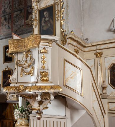 Zdjęcie nr 3: Ambona przyścienna, wisząca, z dostępem od strony prezbiterium w postaci jednobiegowych, zakręconych w górnej partii schodów z pełną balustradą dekorowaną płycinami w kształcie rombów oraz poręczą w formie profilowanego gzymsu. Kosz na planie kwadratu o ściętych narożach i wklęsłych bokach, ustawiony na ćwierćkolistym wałku dekorowanym liśćmi i muszlą; z podwieszeniem w formie ostrosłupa o ściętym wierzchołku i narożach ujętych czterema spływami wolutowymi dekorowanymi akantem i muszlą, zakończonym szyszką. Kosz ujęty w narożach spływami wolutowymi, zdobionymi akantem i kampanulami, podtrzymującymi niepełne belkowanie z silnie wysuniętym gzymsem stanowiącym parapet. W polach ścianek kartusze z podwieszonymi girlandami. W narożu pulpit w postaci postumentu z rzeźbioną księgą. Zaplecek w formie wydłużonego prostokąta flankowanego pilastrami dekorowanymi zwisami, zakończonymi konsolami dźwigającymi imposty z silnie wysuniętym gzymsem oraz fragmenty przerwanego, wolutowego, odwróconego przyczółka. W polu zawieszony obraz Matki Boskiej z Dzieciątkiem. Zaplecek ujęty spływami wolutowymi oraz uszami w postaci strzępionych liści palmowych. Całość zwieńczona glorią promienistą z tablicami Dekalogu w centrum, w otoczeniu obłoków. Struktura polichromowana na sposób imitujący marmoryzację w kolorze beżowym, ornamentyka i profile złocone, detale srebrzone.