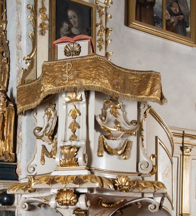 Zdjęcie nr 2: Ambona przyścienna, wisząca, z dostępem od strony prezbiterium w postaci jednobiegowych, zakręconych w górnej partii schodów z pełną balustradą dekorowaną płycinami w kształcie rombów oraz poręczą w formie profilowanego gzymsu. Kosz na planie kwadratu o ściętych narożach i wklęsłych bokach, ustawiony na ćwierćkolistym wałku dekorowanym liśćmi i muszlą; z podwieszeniem w formie ostrosłupa o ściętym wierzchołku i narożach ujętych czterema spływami wolutowymi dekorowanymi akantem i muszlą, zakończonym szyszką. Kosz ujęty w narożach spływami wolutowymi, zdobionymi akantem i kampanulami, podtrzymującymi niepełne belkowanie z silnie wysuniętym gzymsem stanowiącym parapet. W polach ścianek kartusze z podwieszonymi girlandami. W narożu pulpit w postaci postumentu z rzeźbioną księgą. Zaplecek w formie wydłużonego prostokąta flankowanego pilastrami dekorowanymi zwisami, zakończonymi konsolami dźwigającymi imposty z silnie wysuniętym gzymsem oraz fragmenty przerwanego, wolutowego, odwróconego przyczółka. W polu zawieszony obraz Matki Boskiej z Dzieciątkiem. Zaplecek ujęty spływami wolutowymi oraz uszami w postaci strzępionych liści palmowych. Całość zwieńczona glorią promienistą z tablicami Dekalogu w centrum, w otoczeniu obłoków. Struktura polichromowana na sposób imitujący marmoryzację w kolorze beżowym, ornamentyka i profile złocone, detale srebrzone.