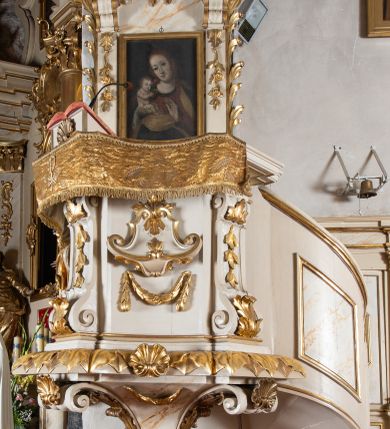 Zdjęcie nr 1: Ambona przyścienna, wisząca, z dostępem od strony prezbiterium w postaci jednobiegowych, zakręconych w górnej partii schodów z pełną balustradą dekorowaną płycinami w kształcie rombów oraz poręczą w formie profilowanego gzymsu. Kosz na planie kwadratu o ściętych narożach i wklęsłych bokach, ustawiony na ćwierćkolistym wałku dekorowanym liśćmi i muszlą; z podwieszeniem w formie ostrosłupa o ściętym wierzchołku i narożach ujętych czterema spływami wolutowymi dekorowanymi akantem i muszlą, zakończonym szyszką. Kosz ujęty w narożach spływami wolutowymi, zdobionymi akantem i kampanulami, podtrzymującymi niepełne belkowanie z silnie wysuniętym gzymsem stanowiącym parapet. W polach ścianek kartusze z podwieszonymi girlandami. W narożu pulpit w postaci postumentu z rzeźbioną księgą. Zaplecek w formie wydłużonego prostokąta flankowanego pilastrami dekorowanymi zwisami, zakończonymi konsolami dźwigającymi imposty z silnie wysuniętym gzymsem oraz fragmenty przerwanego, wolutowego, odwróconego przyczółka. W polu zawieszony obraz Matki Boskiej z Dzieciątkiem. Zaplecek ujęty spływami wolutowymi oraz uszami w postaci strzępionych liści palmowych. Całość zwieńczona glorią promienistą z tablicami Dekalogu w centrum, w otoczeniu obłoków. Struktura polichromowana na sposób imitujący marmoryzację w kolorze beżowym, ornamentyka i profile złocone, detale srebrzone.