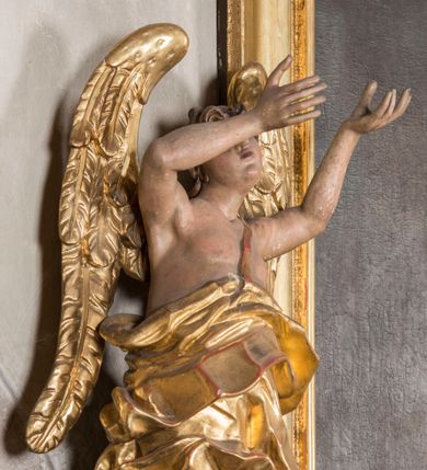 Zdjęcie nr 1: Figura ścięta i drążona z tyłu, ustawiona na niskiej podstawie. Anioł ukazany frontalnie, w kontrapoście, o esowato wygiętej sylwetce, obie ręce unosi wysoko, kierując je w lewą stronę. Twarz ma okrągłą, o pełnych policzkach i wyodrębnionej brodzie, migdałowatych oczach, prostym nosie i rozchylonych ustach, okoloną krótkimi, kręconymi, brązowymi włosami. Anioł ubrany w ekspresyjnie fałdowany, złocony płaszcz, podtrzymywany pojedynczą taśmą przełożoną przez lewe ramię, zawinięty wokół bioder, odsłaniający lewą nogę. Skrzydła ma duże, złożone i złocone. Przy prawej nodze postaci srebrzony obłok. Polichromia ciała naturalistyczna.  



