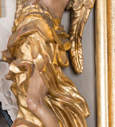 Zdjęcie nr 2: Figura ścięta i drążona z tyłu, ustawiona na niskiej podstawie. Anioł ukazany frontalnie, w kontrapoście, o esowato wygiętej sylwetce, obie ręce unosi wysoko, kierując je w lewą stronę. Twarz ma okrągłą, o pełnych policzkach i wyodrębnionej brodzie, migdałowatych oczach, prostym nosie i rozchylonych ustach, okoloną krótkimi, kręconymi, brązowymi włosami. Anioł ubrany w ekspresyjnie fałdowany, złocony płaszcz, podtrzymywany pojedynczą taśmą przełożoną przez lewe ramię, zawinięty wokół bioder, odsłaniający lewą nogę. Skrzydła ma duże, złożone i złocone. Przy prawej nodze postaci srebrzony obłok. Polichromia ciała naturalistyczna.  



