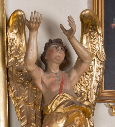 Zdjęcie nr 1: Figura ścięta i drążona z tyłu, ustawiona na niskiej, nieregularnej podstawie. Anioł ukazany frontalnie, z ugiętą prawą nogą opartą na obłoku, o esowato wygiętej sylwetce, z obiema rękami uniesionymi wysoko i skierowanymi w prawo. Twarz ma okrągłą, o pełnych policzkach i wyodrębnionej brodzie, migdałowatych oczach, prostym nosie i rozchylonych ustach, okoloną krótkimi, kręconymi, brązowymi włosami. Anioł ubrany jest w ekspresyjnie fałdowany, złocony płaszcz, podtrzymywany pojedynczą taśmą przełożoną przez lewe ramię, zawinięty wokół bioder i nóg, odsłaniający tors. Skrzydła duże, złożone i złocone. Polichromia ciała naturalistyczna.
