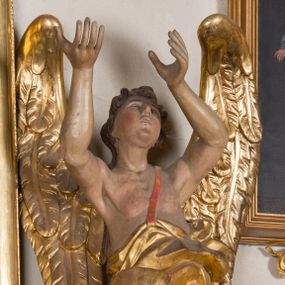 Zdjęcie nr 1: Figura ścięta i drążona z tyłu, ustawiona na niskiej, nieregularnej podstawie. Anioł ukazany frontalnie, z ugiętą prawą nogą opartą na obłoku, o esowato wygiętej sylwetce, z obiema rękami uniesionymi wysoko i skierowanymi w prawo. Twarz ma okrągłą, o pełnych policzkach i wyodrębnionej brodzie, migdałowatych oczach, prostym nosie i rozchylonych ustach, okoloną krótkimi, kręconymi, brązowymi włosami. Anioł ubrany jest w ekspresyjnie fałdowany, złocony płaszcz, podtrzymywany pojedynczą taśmą przełożoną przez lewe ramię, zawinięty wokół bioder i nóg, odsłaniający tors. Skrzydła duże, złożone i złocone. Polichromia ciała naturalistyczna.
