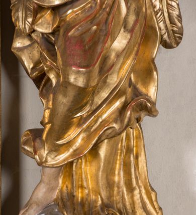 Zdjęcie nr 2: Figura ścięta i drążona z tyłu, ustawiona na niskiej, nieregularnej podstawie. Anioł ukazany frontalnie, z ugiętą prawą nogą opartą na obłoku, o esowato wygiętej sylwetce, z obiema rękami uniesionymi wysoko i skierowanymi w prawo. Twarz ma okrągłą, o pełnych policzkach i wyodrębnionej brodzie, migdałowatych oczach, prostym nosie i rozchylonych ustach, okoloną krótkimi, kręconymi, brązowymi włosami. Anioł ubrany jest w ekspresyjnie fałdowany, złocony płaszcz, podtrzymywany pojedynczą taśmą przełożoną przez lewe ramię, zawinięty wokół bioder i nóg, odsłaniający tors. Skrzydła duże, złożone i złocone. Polichromia ciała naturalistyczna.
