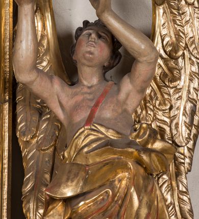 Zdjęcie nr 3: Figura ścięta i drążona z tyłu, ustawiona na niskiej, nieregularnej podstawie. Anioł ukazany frontalnie, z ugiętą prawą nogą opartą na obłoku, o esowato wygiętej sylwetce, z obiema rękami uniesionymi wysoko i skierowanymi w prawo. Twarz ma okrągłą, o pełnych policzkach i wyodrębnionej brodzie, migdałowatych oczach, prostym nosie i rozchylonych ustach, okoloną krótkimi, kręconymi, brązowymi włosami. Anioł ubrany jest w ekspresyjnie fałdowany, złocony płaszcz, podtrzymywany pojedynczą taśmą przełożoną przez lewe ramię, zawinięty wokół bioder i nóg, odsłaniający tors. Skrzydła duże, złożone i złocone. Polichromia ciała naturalistyczna.
