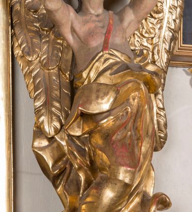 Zdjęcie nr 5: Figura ścięta i drążona z tyłu, ustawiona na niskiej, nieregularnej podstawie. Anioł ukazany frontalnie, z ugiętą prawą nogą opartą na obłoku, o esowato wygiętej sylwetce, z obiema rękami uniesionymi wysoko i skierowanymi w prawo. Twarz ma okrągłą, o pełnych policzkach i wyodrębnionej brodzie, migdałowatych oczach, prostym nosie i rozchylonych ustach, okoloną krótkimi, kręconymi, brązowymi włosami. Anioł ubrany jest w ekspresyjnie fałdowany, złocony płaszcz, podtrzymywany pojedynczą taśmą przełożoną przez lewe ramię, zawinięty wokół bioder i nóg, odsłaniający tors. Skrzydła duże, złożone i złocone. Polichromia ciała naturalistyczna.
