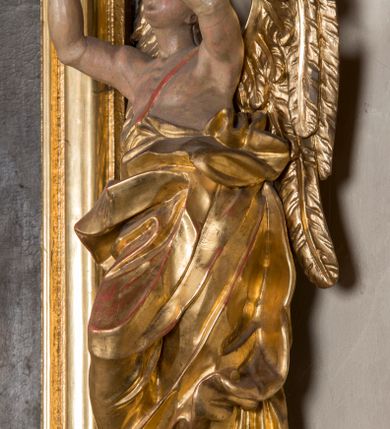 Zdjęcie nr 6: Figura ścięta i drążona z tyłu, ustawiona na niskiej, nieregularnej podstawie. Anioł ukazany frontalnie, z ugiętą prawą nogą opartą na obłoku, o esowato wygiętej sylwetce, z obiema rękami uniesionymi wysoko i skierowanymi w prawo. Twarz ma okrągłą, o pełnych policzkach i wyodrębnionej brodzie, migdałowatych oczach, prostym nosie i rozchylonych ustach, okoloną krótkimi, kręconymi, brązowymi włosami. Anioł ubrany jest w ekspresyjnie fałdowany, złocony płaszcz, podtrzymywany pojedynczą taśmą przełożoną przez lewe ramię, zawinięty wokół bioder i nóg, odsłaniający tors. Skrzydła duże, złożone i złocone. Polichromia ciała naturalistyczna.
