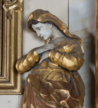 Zdjęcie nr 1: Rzeźba ścięta z tyłu, głęboko drążona, ustawiona na ćwierćkolistej podstawie. Matka Boska ukazana frontalnie, w kontrapoście, o esowato wygiętej sylwetce, odchylonym delikatnie w tył korpusie, z silnie pochyloną głową. Ręce ma skrzyżowane i złożone na piersi. Twarz ma owalną, o delikatnych, młodzieńczych rysach, małych oczach, skierowanych w dół, dużym nosie oraz niewielkich ustach. Włosy ma długie, falowane, z przedziałkiem na środku głowy, schowane pod welonem. Maria ubrana jest w tunikę, z długimi, wąskimi rękawami, przepasaną w talii, ściśle przylegającą do ciała i marszczoną w partii klatki piersiowej. Na głowę ma narzucony silnie drapowany płaszcz, opadający na plecy i zarzucony z przodu postaci, z wywiniętym fragmentem. Polichromia naturalistyczna w odsłoniętych partiach ciała, szaty złocone.

