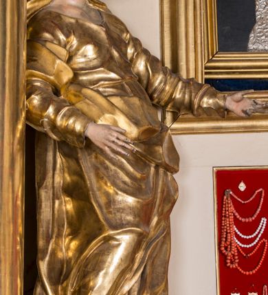 Zdjęcie nr 6: Rzeźba ścięta z tyłu, głęboko drążona, ustawiona na ćwierćkolistej podstawie. Figura ukazana frontalnie, w kontrapoście, o esowato wygiętej sylwetce, przechylonej delikatnie w tył, z głową skierowaną w lewo. Obie ręce ma ugięte i wyciągnięte do przodu. Twarz ma owalną, o delikatnych, młodzieńczych rysach, małych oczach, dużym nosie i niewielkich ustach. Włosy ma długie, falowane, z przedziałkiem na środku głowy, schowane pod welonem. Święta ubrana jest w tunikę, z długimi, wąskimi rękawami, przepasaną w talii, ściśle przylegającą do ciała. Na głowę ma narzucony silnie drapowany płaszcz, opadający na plecy, zawiązany na prawym ramieniu i przerzucony przez biodra z wywiniętym z przodu brzegiem. Polichromia naturalistyczna w partiach ciała, szaty złocone.