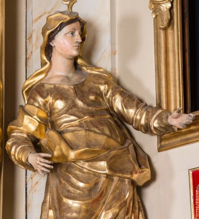 Zdjęcie nr 1: Rzeźba ścięta z tyłu, głęboko drążona, ustawiona na ćwierćkolistej podstawie. Figura ukazana frontalnie, w kontrapoście, o esowato wygiętej sylwetce, przechylonej delikatnie w tył, z głową skierowaną w lewo. Obie ręce ma ugięte i wyciągnięte do przodu. Twarz ma owalną, o delikatnych, młodzieńczych rysach, małych oczach, dużym nosie i niewielkich ustach. Włosy ma długie, falowane, z przedziałkiem na środku głowy, schowane pod welonem. Święta ubrana jest w tunikę, z długimi, wąskimi rękawami, przepasaną w talii, ściśle przylegającą do ciała. Na głowę ma narzucony silnie drapowany płaszcz, opadający na plecy, zawiązany na prawym ramieniu i przerzucony przez biodra z wywiniętym z przodu brzegiem. Polichromia naturalistyczna w partiach ciała, szaty złocone.