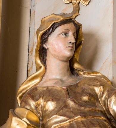 Zdjęcie nr 4: Rzeźba ścięta z tyłu, głęboko drążona, ustawiona na ćwierćkolistej podstawie. Figura ukazana frontalnie, w kontrapoście, o esowato wygiętej sylwetce, przechylonej delikatnie w tył, z głową skierowaną w lewo. Obie ręce ma ugięte i wyciągnięte do przodu. Twarz ma owalną, o delikatnych, młodzieńczych rysach, małych oczach, dużym nosie i niewielkich ustach. Włosy ma długie, falowane, z przedziałkiem na środku głowy, schowane pod welonem. Święta ubrana jest w tunikę, z długimi, wąskimi rękawami, przepasaną w talii, ściśle przylegającą do ciała. Na głowę ma narzucony silnie drapowany płaszcz, opadający na plecy, zawiązany na prawym ramieniu i przerzucony przez biodra z wywiniętym z przodu brzegiem. Polichromia naturalistyczna w partiach ciała, szaty złocone.