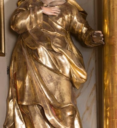 Zdjęcie nr 6: Rzeźba ścięta z tyłu, głęboko drążona, ustawiona na ćwierćkolistej podstawie. Figura ukazana frontalnie, w kontrapoście, o esowato wygiętej sylwetce, delikatnie odchylonej, z nieznacznie uniesioną głową, skierowaną w prawo. Prawą rękę kładzie na piersi, lewą, ugiętą wyciąga przed siebie. Twarz ma owalną, o delikatnych, młodzieńczych rysach, małych oczach, dużym nosie i niewielkich ustach. Włosy ma falowane, z przedziałkiem na środku głowy, ukryte pod welonem. Święta ubrana jest w tunikę, z długimi, wąskimi rękawami, przepasaną w talii, ściśle przylegającą do ciała oraz płaszcz zawieszony w partii nóg, z wywiniętą fałdą na lewym boku. Na głowę ma narzucony welon, opadający na ramiona. Polichromia naturalistyczna w partiach ciała, szaty złocone.