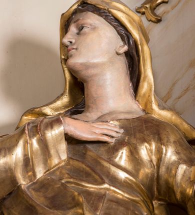 Zdjęcie nr 5: Rzeźba ścięta z tyłu, głęboko drążona, ustawiona na ćwierćkolistej podstawie. Figura ukazana frontalnie, w kontrapoście, o esowato wygiętej sylwetce, delikatnie odchylonej, z nieznacznie uniesioną głową, skierowaną w prawo. Prawą rękę kładzie na piersi, lewą, ugiętą wyciąga przed siebie. Twarz ma owalną, o delikatnych, młodzieńczych rysach, małych oczach, dużym nosie i niewielkich ustach. Włosy ma falowane, z przedziałkiem na środku głowy, ukryte pod welonem. Święta ubrana jest w tunikę, z długimi, wąskimi rękawami, przepasaną w talii, ściśle przylegającą do ciała oraz płaszcz zawieszony w partii nóg, z wywiniętą fałdą na lewym boku. Na głowę ma narzucony welon, opadający na ramiona. Polichromia naturalistyczna w partiach ciała, szaty złocone.