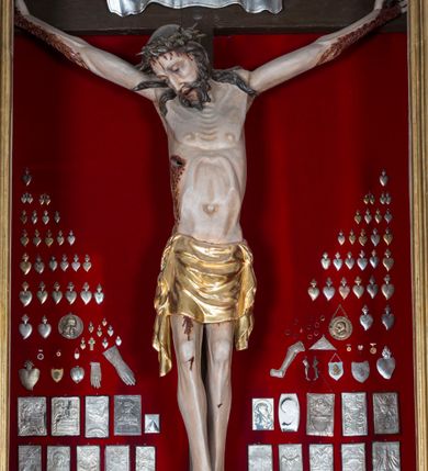 Zdjęcie nr 7: Figura Chrystusa żywego, ścięta z tyłu, przybita do krzyża trzema gwoździami. Ciało w lekkim zwisie, o ramionach układających się na kształt łuku, głowa opadająca na prawy bark, nogi wyprostowane, stopy skrzyżowane w układzie prawa na lewą. Chrystus ma pociągłą twarz, o wystających kościach policzkowych, dużych, półprzymkniętych oczach, długim, prostym nosie oraz szerokich, otwartych ustach z widoczną linią zębów. Twarz okolona jest krótką brodą silnie skręconą w pojedyncze pukle oraz falowanymi włosami opadającymi pojedynczymi pasmami na ramiona. Na głowie ma koronę cierniową. Jego ciało jest smukłe i wychudzone z podkreślonymi żebrami i mostkiem klatki piersiowej oraz ścięgnami w partii ramion i nóg. Stopy i dłonie ma nieproporcjonalnie duże. Prawy bok ma przebity, z widocznym dużym otworem oraz rzeźbioną, obficie broczącą krwią ze wszystkich ran. Perizonium sięgające połowy ud, silnie fałdowane, z dwoma kaskadowymi zwisami tkaniny na obu bokach. Ciało polichromowane naturalistycznie, o bladej karnacji, włosy i broda ciemnobrązowe, perizonium złocone. Na zakończeniu pionowej belki krzyża pozioma, srebrzona banderola z napisem „INRI”. 

