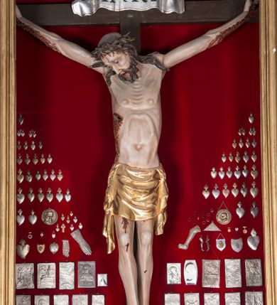 Zdjęcie nr 1: Figura Chrystusa żywego, ścięta z tyłu, przybita do krzyża trzema gwoździami. Ciało w lekkim zwisie, o ramionach układających się na kształt łuku, głowa opadająca na prawy bark, nogi wyprostowane, stopy skrzyżowane w układzie prawa na lewą. Chrystus ma pociągłą twarz, o wystających kościach policzkowych, dużych, półprzymkniętych oczach, długim, prostym nosie oraz szerokich, otwartych ustach z widoczną linią zębów. Twarz okolona jest krótką brodą silnie skręconą w pojedyncze pukle oraz falowanymi włosami opadającymi pojedynczymi pasmami na ramiona. Na głowie ma koronę cierniową. Jego ciało jest smukłe i wychudzone z podkreślonymi żebrami i mostkiem klatki piersiowej oraz ścięgnami w partii ramion i nóg. Stopy i dłonie ma nieproporcjonalnie duże. Prawy bok ma przebity, z widocznym dużym otworem oraz rzeźbioną, obficie broczącą krwią ze wszystkich ran. Perizonium sięgające połowy ud, silnie fałdowane, z dwoma kaskadowymi zwisami tkaniny na obu bokach. Ciało polichromowane naturalistycznie, o bladej karnacji, włosy i broda ciemnobrązowe, perizonium złocone. Na zakończeniu pionowej belki krzyża pozioma, srebrzona banderola z napisem „INRI”. 

