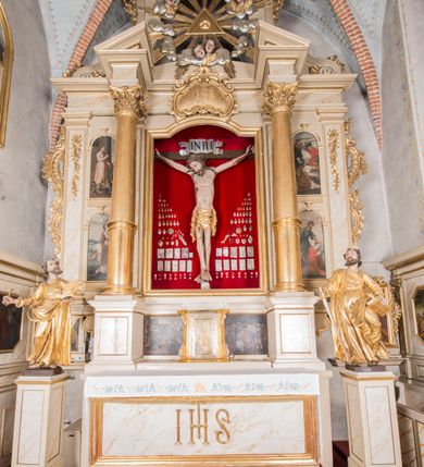 Zdjęcie nr 6: Figura Chrystusa żywego, ścięta z tyłu, przybita do krzyża trzema gwoździami. Ciało w lekkim zwisie, o ramionach układających się na kształt łuku, głowa opadająca na prawy bark, nogi wyprostowane, stopy skrzyżowane w układzie prawa na lewą. Chrystus ma pociągłą twarz, o wystających kościach policzkowych, dużych, półprzymkniętych oczach, długim, prostym nosie oraz szerokich, otwartych ustach z widoczną linią zębów. Twarz okolona jest krótką brodą silnie skręconą w pojedyncze pukle oraz falowanymi włosami opadającymi pojedynczymi pasmami na ramiona. Na głowie ma koronę cierniową. Jego ciało jest smukłe i wychudzone z podkreślonymi żebrami i mostkiem klatki piersiowej oraz ścięgnami w partii ramion i nóg. Stopy i dłonie ma nieproporcjonalnie duże. Prawy bok ma przebity, z widocznym dużym otworem oraz rzeźbioną, obficie broczącą krwią ze wszystkich ran. Perizonium sięgające połowy ud, silnie fałdowane, z dwoma kaskadowymi zwisami tkaniny na obu bokach. Ciało polichromowane naturalistycznie, o bladej karnacji, włosy i broda ciemnobrązowe, perizonium złocone. Na zakończeniu pionowej belki krzyża pozioma, srebrzona banderola z napisem „INRI”. 

