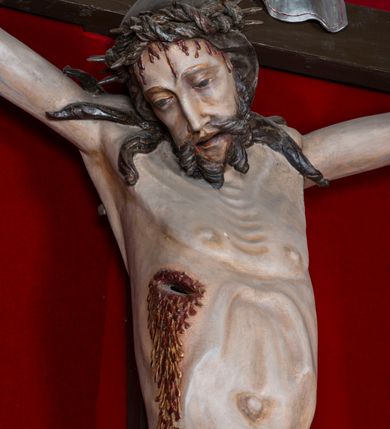 Zdjęcie nr 2: Figura Chrystusa żywego, ścięta z tyłu, przybita do krzyża trzema gwoździami. Ciało w lekkim zwisie, o ramionach układających się na kształt łuku, głowa opadająca na prawy bark, nogi wyprostowane, stopy skrzyżowane w układzie prawa na lewą. Chrystus ma pociągłą twarz, o wystających kościach policzkowych, dużych, półprzymkniętych oczach, długim, prostym nosie oraz szerokich, otwartych ustach z widoczną linią zębów. Twarz okolona jest krótką brodą silnie skręconą w pojedyncze pukle oraz falowanymi włosami opadającymi pojedynczymi pasmami na ramiona. Na głowie ma koronę cierniową. Jego ciało jest smukłe i wychudzone z podkreślonymi żebrami i mostkiem klatki piersiowej oraz ścięgnami w partii ramion i nóg. Stopy i dłonie ma nieproporcjonalnie duże. Prawy bok ma przebity, z widocznym dużym otworem oraz rzeźbioną, obficie broczącą krwią ze wszystkich ran. Perizonium sięgające połowy ud, silnie fałdowane, z dwoma kaskadowymi zwisami tkaniny na obu bokach. Ciało polichromowane naturalistycznie, o bladej karnacji, włosy i broda ciemnobrązowe, perizonium złocone. Na zakończeniu pionowej belki krzyża pozioma, srebrzona banderola z napisem „INRI”. 

