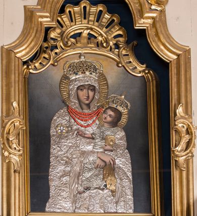 Zdjęcie nr 4: Obraz w formie stojącego prostokąta zamkniętego łukiem półkolistym nadwieszonym z uskokiem, ujęty profilowaną, złoconą ramą dekorowaną u szczytu rocaille'em. Partie ciała postaci malowane, ubiór zakryty srebrzonymi sukienkami dekorowanymi trybowanym wzorem roślinnym. W centrum półpostać Matki Boskiej zwróconej delikatnie w lewo, podtrzymującej na lewym ramieniu Dzieciątko, ze skrzyżowanymi dłońmi, trzymającymi chustkę. Twarz ma pociągłą, o trójkątnie zarysowanej brodzie oraz silnie podkreślonych światłocieniem liniach łuków brwiowych i nosa, o migdałowatych oczach, prostym nosie i niewielkich, pełnych ustach. Ubrana jest w maforion narzucony na głowę i ramiona, na prawym barku ma kameryzowaną gwiazdę. Wokół szyi ma trzy sznury korali. Dzieciątko siedzi na ramieniu Matki, zwrócone  w trzech czwartych w prawo, z uniesioną głową. W lewej ręce trzyma przy boku księgę, prawą wyciąga w geście błogosławieństwa. Twarz ma pełną, o trójkątnej linii brody, oczach zwróconych ku górze, prostym nosie i pełnych ustach; okoloną krótkimi, silnie kręconymi włosami. Ubrane jest w luźną tunikę, na stopach ma sandały. Na głowach obu postaci nałożone metalowe, złocone i kameryzowane korony zamknięte, wokół nich metalowe i złocone nimby zamknięte. Tło cieniowane, niebiesko-szare, rozświetlone nad głową Marii. 

